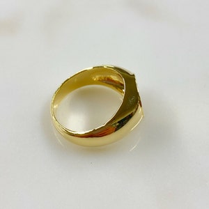 May include: A gold-colored ring with a rounded rectangular face. The ring is highly polished, reflecting light, and appears to be made of solid metal. The band is smooth and wide, with a classic design.