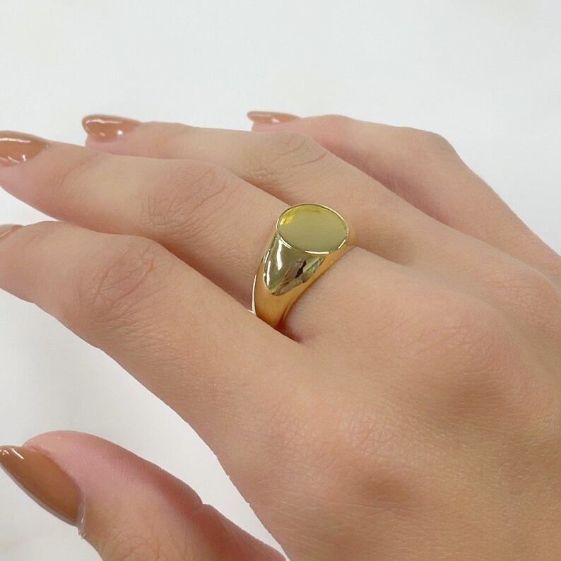 May include: A gold signet ring with a smooth, oval face. The ring is a bright, polished gold color and is worn on a finger. The ring's design is simple and classic, suitable for everyday wear. Jewelry.