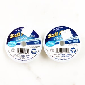 Op de afbeelding: Twee spoelen Soft Flex kralendraad, een met het label "Fijn" en de andere met "Medium". Beide spoelen zijn satijn zilver van kleur en hebben een treksterkte van respectievelijk 10 lbs en 26 lbs. De spoelen zijn gelabeld met de diameter en lengte van de draad in zowel inches als millimeters.