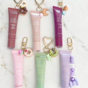 May include: Assortment of Laneige lip gloss tubes in various colors, each attached to a gold keychain with a charm. Flavors include cinnamon sugar, sugar, blueberry, strawberry, matcha, and taro. The tubes are approximately 3 inches long.