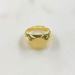 May include: A gold signet ring with a polished, rounded band and a flat, circular face. The ring is a bright, shiny gold color and appears to be made of metal. The ring is a classic style, suitable for everyday wear.