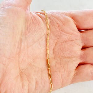 K&ouml;nnte beinhalten: Eine goldene Kettenkette mit einem zarten, d&uuml;nnen und flachen Glieder-Stil. Die Kette wird auf der Hand einer Person gezeigt.