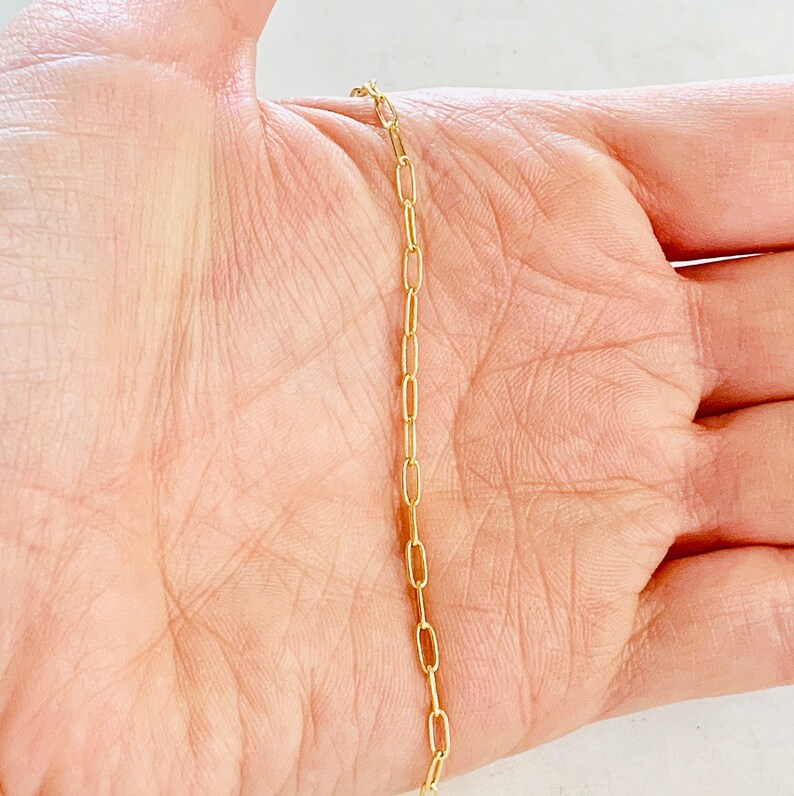 K&ouml;nnte beinhalten: Eine goldene Kettenkette mit einem zarten, d&uuml;nnen und flachen Glieder-Stil. Die Kette wird auf der Hand einer Person gezeigt.