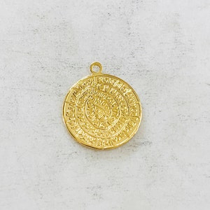 May include: Gold-toned pendant with a circular design featuring intricate patterns and symbols. The pendant has a loop at the top for attaching to a chain.