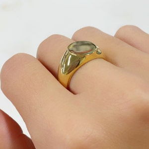 May include: A gold signet ring with a polished finish. The ring features a smooth, oval-shaped face and a thick band. The ring is shown on a person's finger.