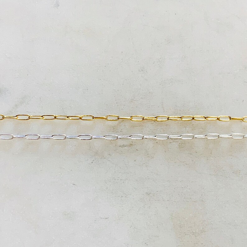 K&ouml;nnte beinhalten: Zwei Ketten, eine goldene und eine silberne, liegen auf einer wei&szlig;en Oberfl&auml;che. Beide Ketten bestehen aus kleinen, ovalen Gliedern.