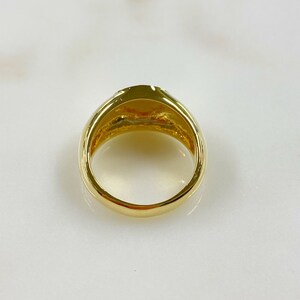 May include: A gold ring with a rounded band and a flat, oval top. The ring has a polished finish, reflecting light. The design is simple and elegant, suitable for everyday wear or special occasions. The ring is a classic piece of jewelry.