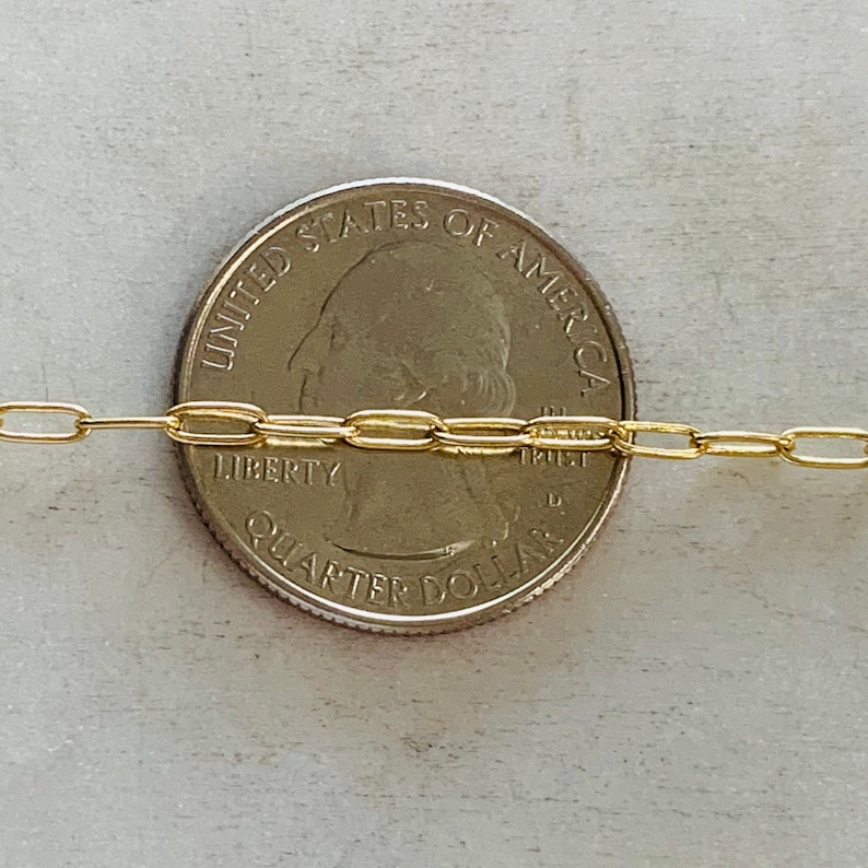 K&ouml;nnte beinhalten: Eine goldene Kettenkette mit ovalen Gliedern, die &uuml;ber eine US-amerikanischen 25-Cent-M&uuml;nze gelegt wird. Die M&uuml;nze zeigt die Vorderseite mit dem Profil von George Washington und den Worten "United States of America", "Liberty" und "In God We Trust".