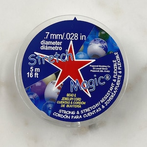 Könnte beinhalten: Eine Spule aus klarem, elastischem Stretch-Perlen- und Schmuckband, genannt "Stretch Magic". Die Spule ist mit dem Text "Stretch Magic" und "Strong & Stretchy/Resistant & Flexible" beschriftet. Die Spule ist 5 Meter oder 16 Fuß lang und das Band hat einen Durchmesser von 0,7 mm oder 0,028 Zoll.