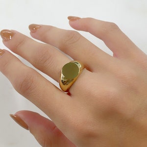 May include: A gold-colored signet ring with a smooth, circular face. The ring has a polished finish and a thick band. The ring is worn on a finger.