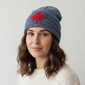 May include: A blue knit beanie featuring a red maple leaf design. The hat is made of a soft, textured material and is worn by a person. The beanie is a stylish accessory for cold weather.