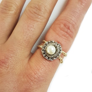 Puede incluir: Primer plano de un anillo con una perla central rodeada por un diseño de filigrana plateada y dorada. El anillo está en un dedo, mostrando los intrincados detalles de la joya.
