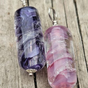 May include: Two glass pendants with silver-colored findings. One pendant is filled with swirling shades of purple and white. The other pendant has pink and white swirls. Both have a cylindrical shape and are displayed on a wooden surface.