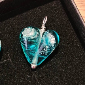 May include: A teal blue glass heart pendant with silver accents. The heart has a swirl pattern and is attached to a silver bail.