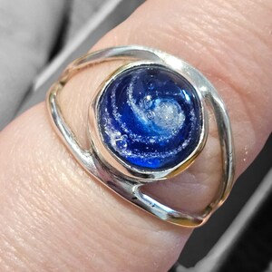 May include: A silver ring with a blue and white galaxy-like design in the center. The ring is on a finger.