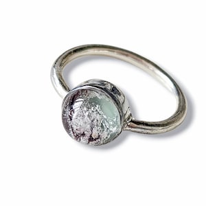 May include: A silver ring with a round, clear glass stone containing a mixture of white, gray, and black particles.