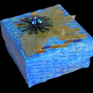 May include: A blue decorative box with a textured surface and a floral embellishment. The box features a geometric pattern with gold and blue accents. The top is adorned with a gold leaf and a dark green and blue flower.