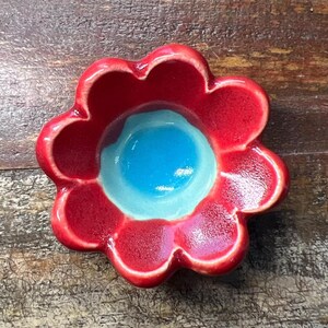 May include: A small ceramic dish shaped like a flower with eight rounded petals. The petals are finished in a glossy red glaze, while the center of the dish features a smooth, circular basin with a vibrant, bright blue glaze finish.