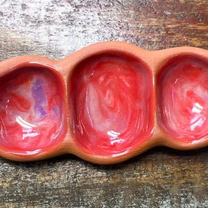 May include: A terracotta-coloured, three-section serving dish. Each section is a rounded bowl with a glossy, swirled red and purple interior. The dish is designed for serving dips, sauces, or small portions of food.