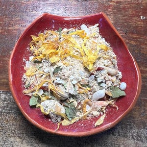 May include: A glossy red ceramic bowl containing a dry mixture of beige powder, scattered yellow flower petals, small green leaves, and white crystalline chunks. The bowl has a distinct curved rim and a smooth, reflective surface texture.