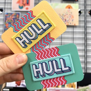 May include: Two colourful iron-on patches with the word "HULL" in white letters on a yellow and turquoise background. The patches have a zig-zag pattern around the edges.
