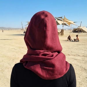 Könnte beinhalten: Eine tiefburgunderrote Kapuze aus strukturiertem Stoff, getragen über einem schwarzen Langarmshirt. Die Kapuze zeichnet sich durch ein drapiertes, locker sitzendes Design aus, das Kopf und Hals bedeckt und eine geschichtete Optik erzeugt.