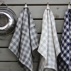 Op de afbeelding: Drie geruite keukendoeken hangen aan haken. De doeken zijn in blauw en wit, beige en wit en donkerblauw en wit.