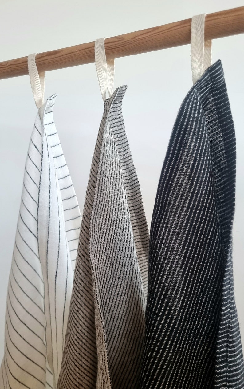 May include: Three striped kitchen towels hanging on a wooden rod. The towels are white with black stripes, beige with black stripes, and black with white stripes.