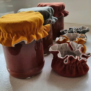 May include: Four glass jars with brown liquid inside, topped with fabric lids in various colors: mustard yellow, green, and brown. The lids have a ruffled edge and are made of a soft, textured fabric.