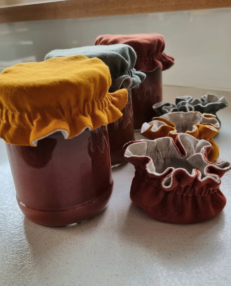 May include: Four glass jars with brown liquid inside, topped with fabric lids in various colors: mustard yellow, green, and brown. The lids have a ruffled edge and are made of a soft, textured fabric.