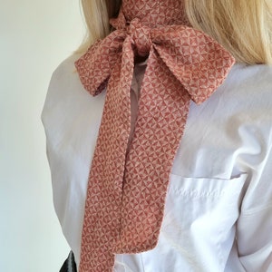 May include: A red and white patterned bow tie worn around the neck. The bow tie is made of a woven fabric with a geometric design.