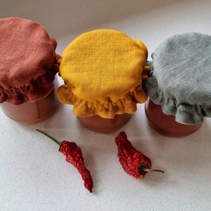 May include: Three glass jars with red lids are topped with fabric covers in rust, yellow, and gray. Two dried red chili peppers are in the foreground.