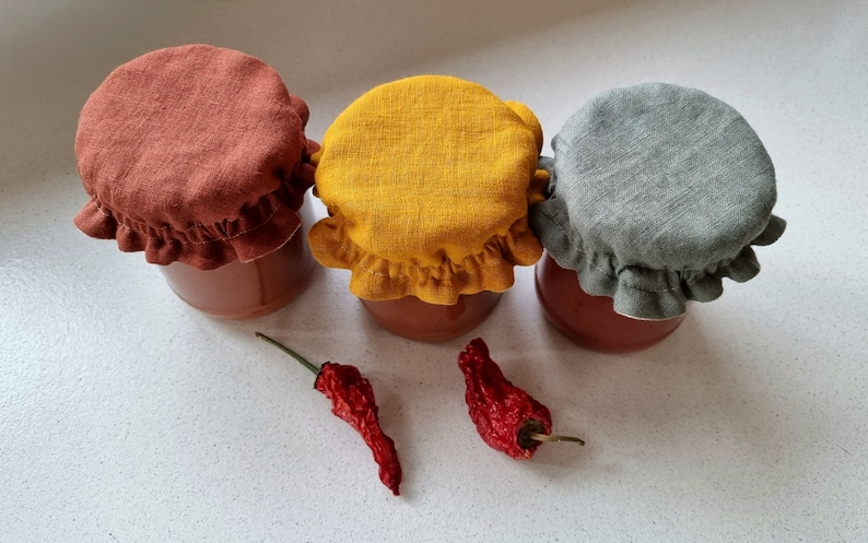 May include: Three glass jars with red lids are topped with fabric covers in rust, yellow, and gray. Two dried red chili peppers are in the foreground.