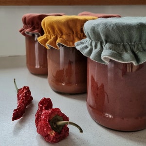 May include: Four glass jars filled with a red sauce, topped with fabric lids in different colors: brown, yellow, green, and white. Two dried red chili peppers are in the foreground.