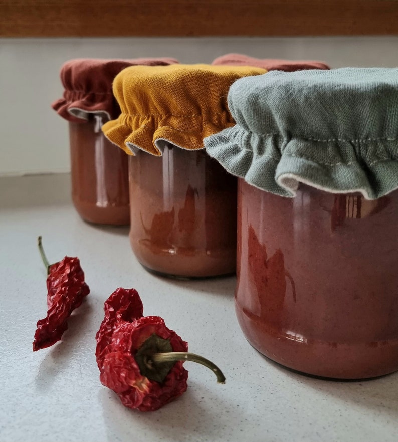 May include: Four glass jars filled with a red sauce, topped with fabric lids in different colors: brown, yellow, green, and white. Two dried red chili peppers are in the foreground.