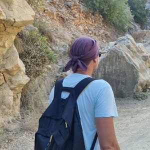 May include: A person wearing a purple bandana and a light blue t-shirt is carrying a black backpack with yellow accents. The person is walking on a path in a rocky area.