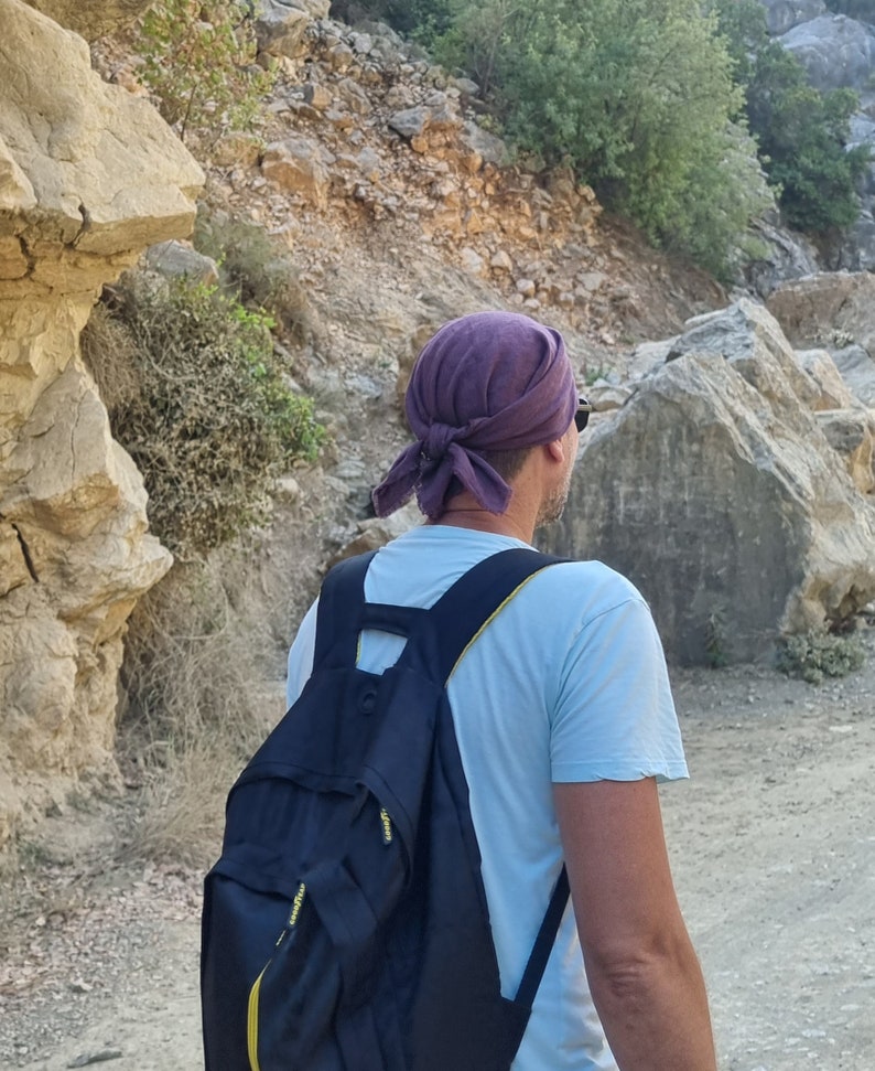May include: A person wearing a purple bandana and a light blue t-shirt is carrying a black backpack with yellow accents. The person is walking on a path in a rocky area.