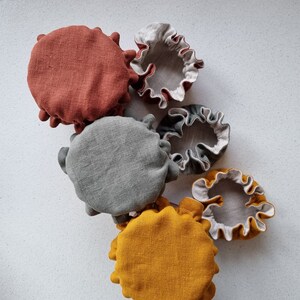 May include: Five fabric food covers in various colors and sizes. The covers are made of a natural linen material and have a ruffled edge. The colors are rust, gray, yellow, and white.