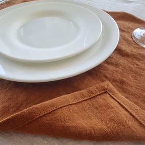 May include: A brown linen placemat with a white plate on top. The placemat is folded over on itself, showing the stitching on the edge.