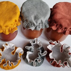 May include: Six fabric jar covers in three colors: mustard yellow, gray, and rust. Three covers are on jars, and three are laying flat. The covers have a ruffled edge.