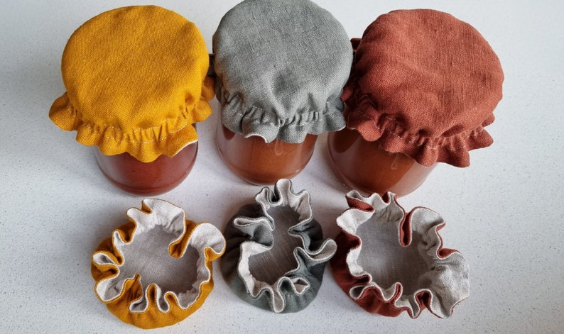 May include: Six fabric jar covers in three colors: mustard yellow, gray, and rust. Three covers are on jars, and three are laying flat. The covers have a ruffled edge.