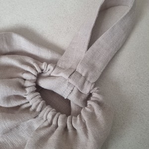 May include: A beige linen bag with a gathered top and two straps. The straps are sewn together at the top of the bag.