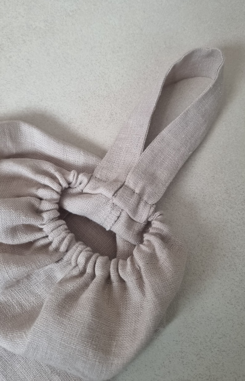 May include: A beige linen bag with a gathered top and two straps. The straps are sewn together at the top of the bag.
