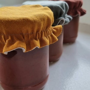 May include: Three glass jars with fabric lids in yellow, gray, and brown. The jars are filled with a dark red liquid.
