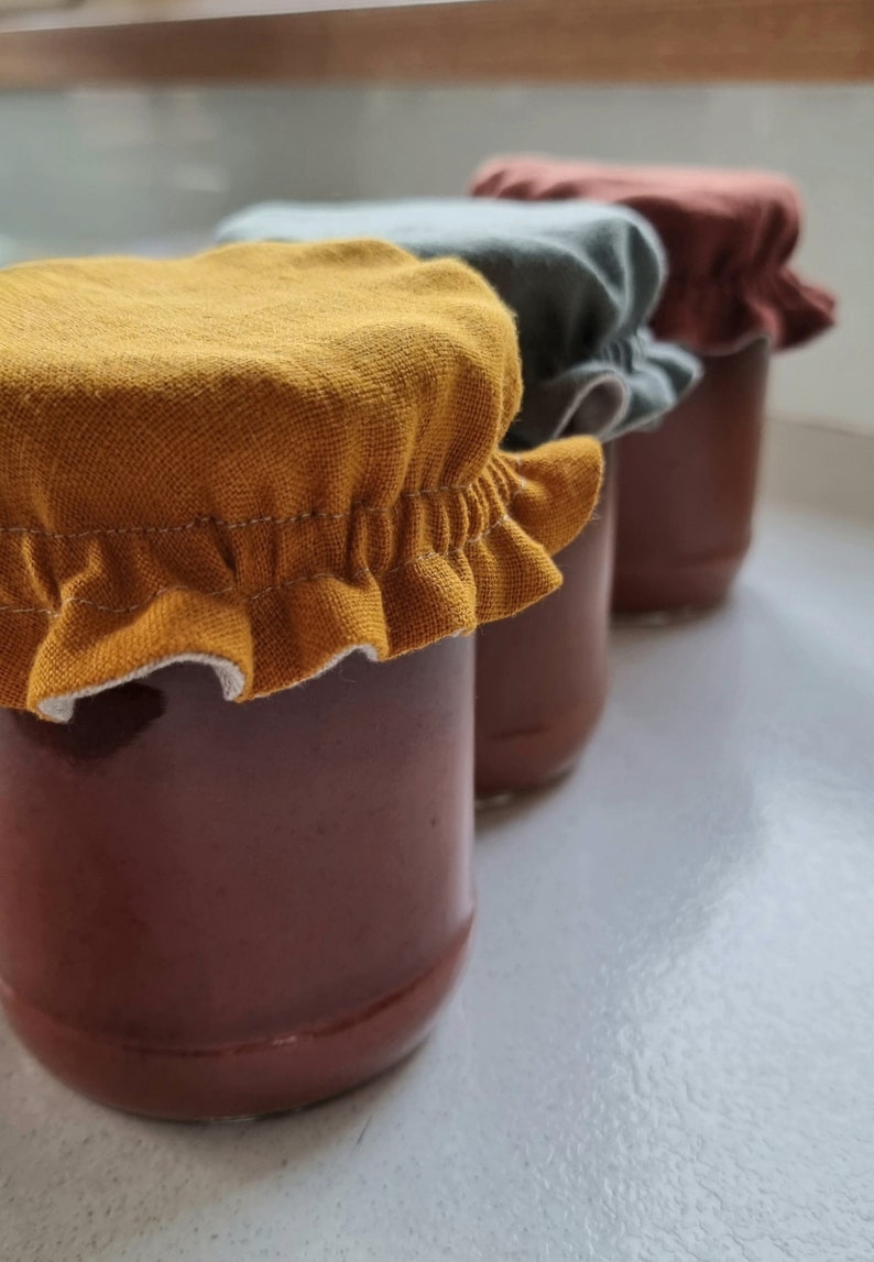 May include: Three glass jars with fabric lids in yellow, gray, and brown. The jars are filled with a dark red liquid.