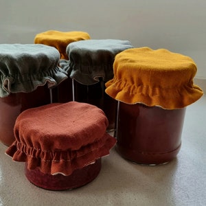 May include: Five glass jars with fabric lids. The lids are in three colors: mustard yellow, forest green, and burnt orange. The jars are filled with a dark red liquid.