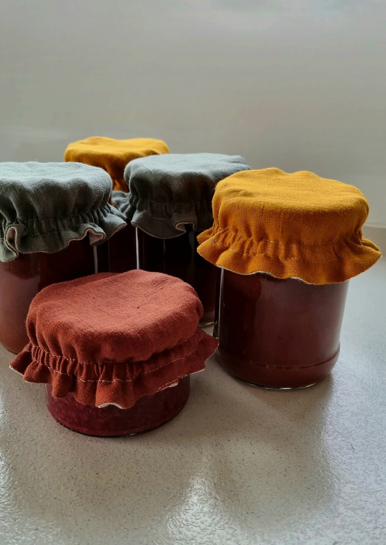 May include: Five glass jars with fabric lids. The lids are in three colors: mustard yellow, forest green, and burnt orange. The jars are filled with a dark red liquid.