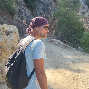 May include: A man wearing a dark purple bandana, sunglasses, and a light blue t-shirt is walking on a dirt path. He is carrying a black backpack with yellow accents.