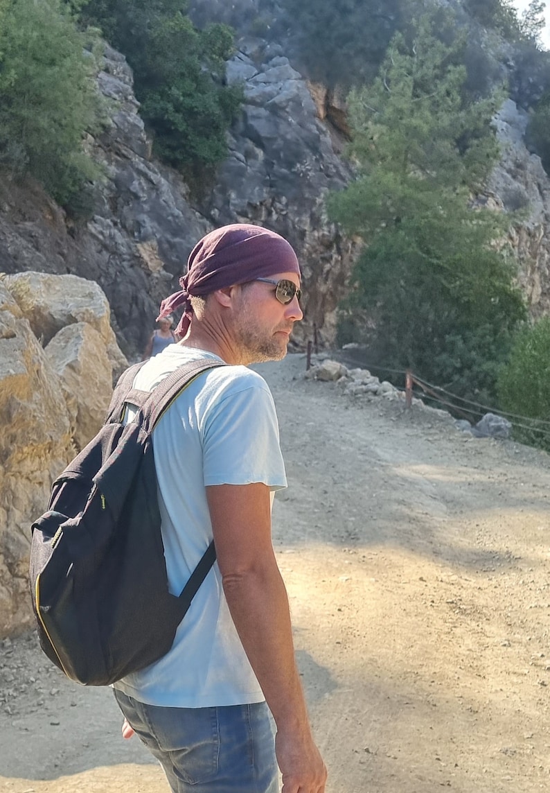 May include: A man wearing a dark purple bandana, sunglasses, and a light blue t-shirt is walking on a dirt path. He is carrying a black backpack with yellow accents.