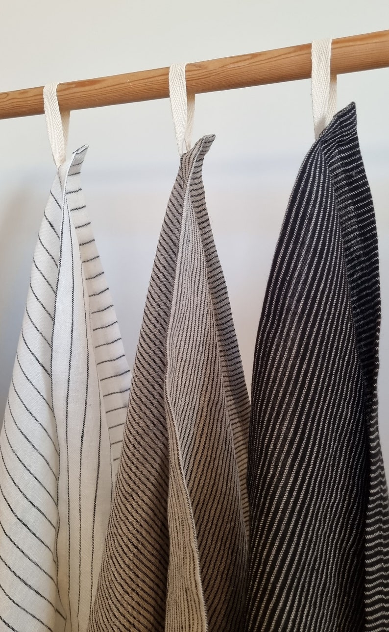 May include: Three striped kitchen towels hanging on a wooden rod. The towels are white with black stripes, beige with black stripes, and black with white stripes.